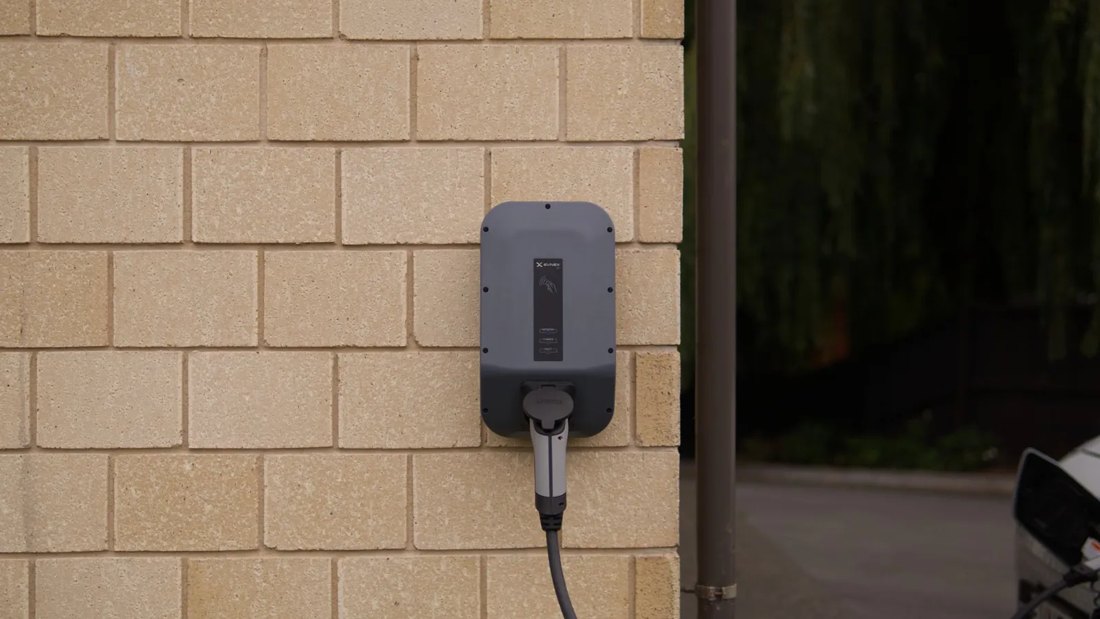 A wall-mounted AC charge point on the outside wall of a building, cable plugged into a car parked alongside
